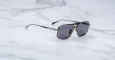A pair of Jacques Marie Mage Earl sunglasses with black frames and dark lenses rests on a white marble surface.