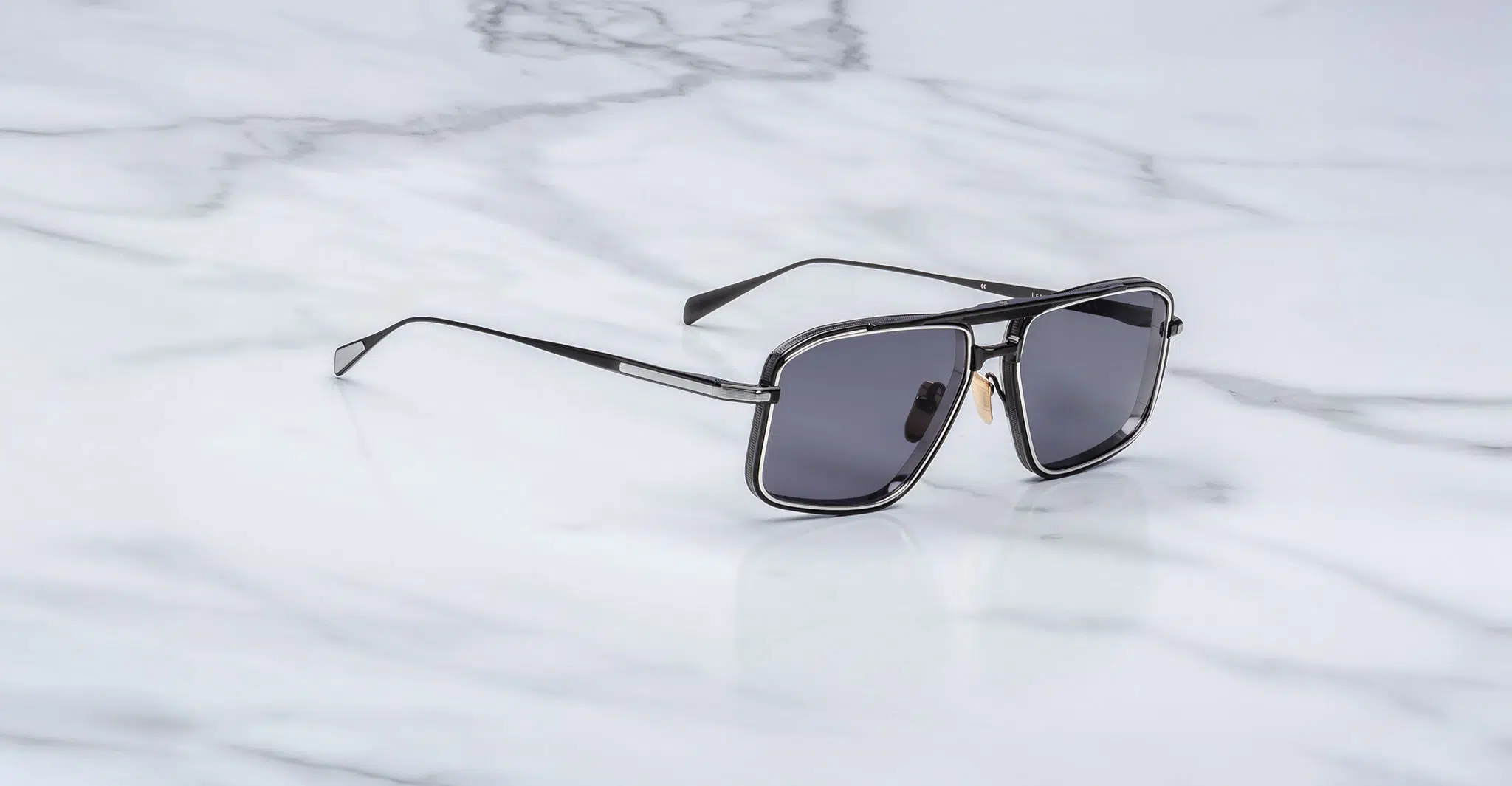 Jacques-Marie-Mage-Earl-Corbeau-JMMER-19J-3-4-Dan-Deutsch A pair of Jacques Marie Mage Earl sunglasses with black frames and dark lenses rests on a white marble surface.