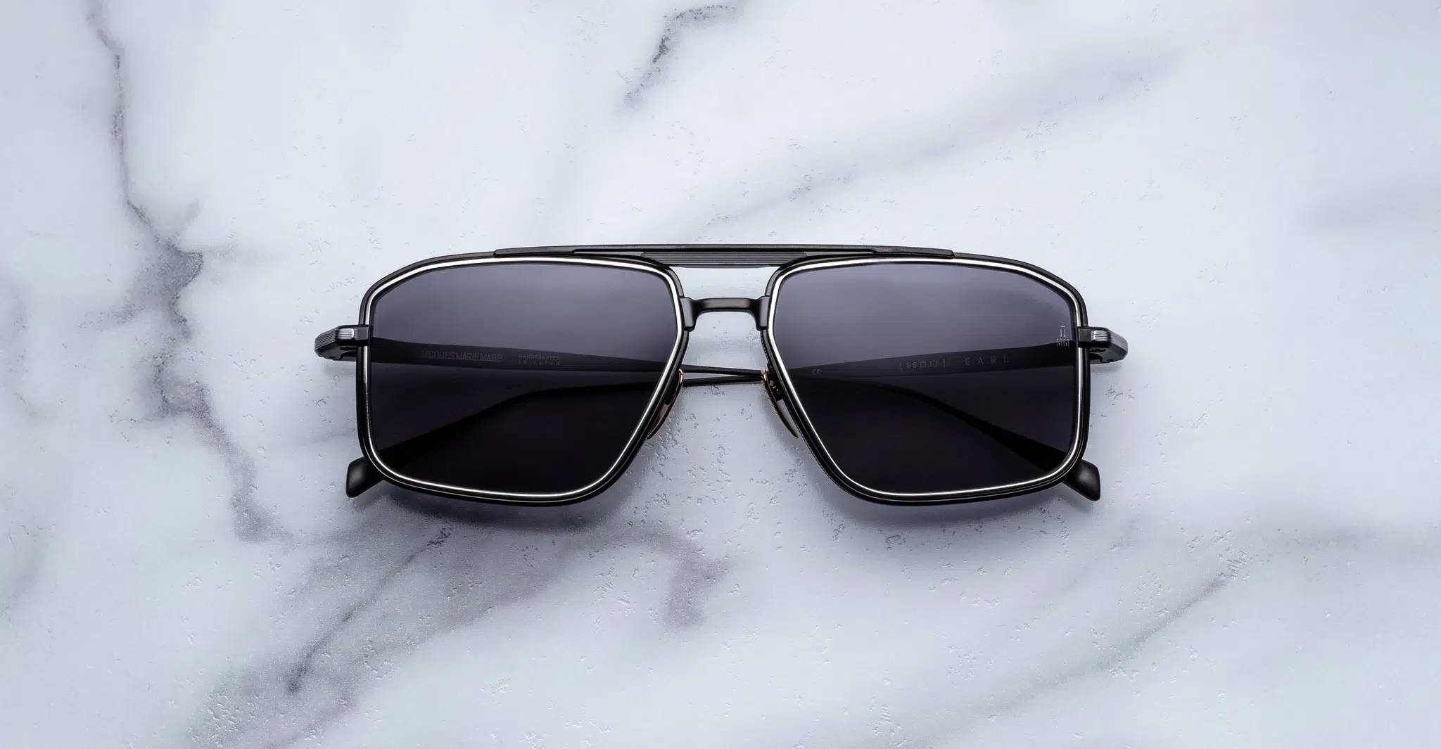 Jacques-Marie-Mage-Earl-Corbeau-JMMER-19J-Front-Dan-Deutsch A pair of Jacques Marie Mage Earl rectangular black sunglasses with dark lenses and a metallic frame is displayed on a white marble surface.