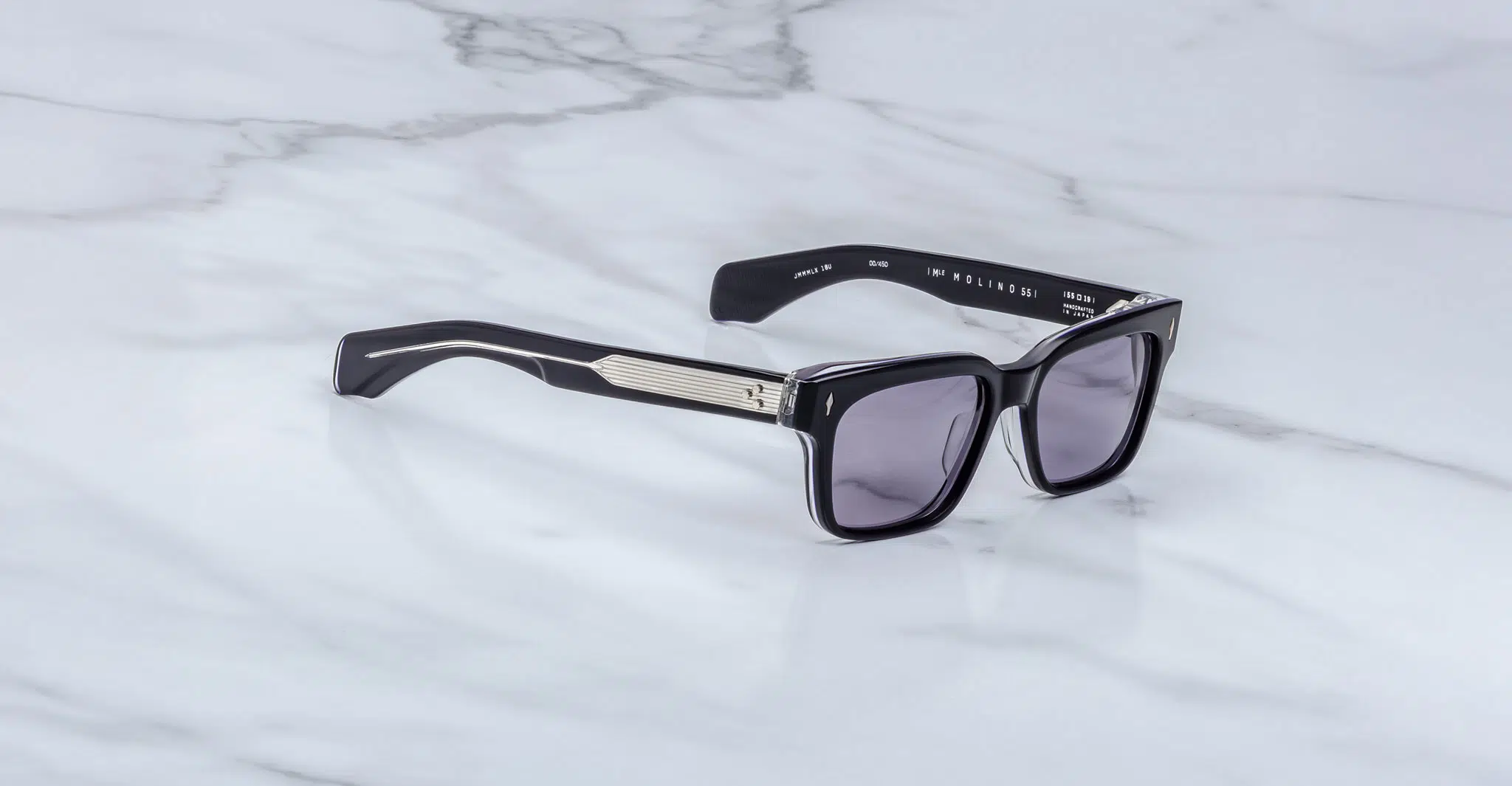 The Jacques Marie Mage Molino 55, featuring black thick frames, silver temple accents, and dark tinted lenses, is displayed on a white marble surface.