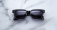 The Jacques Marie Mage Molino 55, black rectangular smart sunglasses with thick frames, rest on a white marble surface. Digital menu interface reflections are visible in their lenses.
