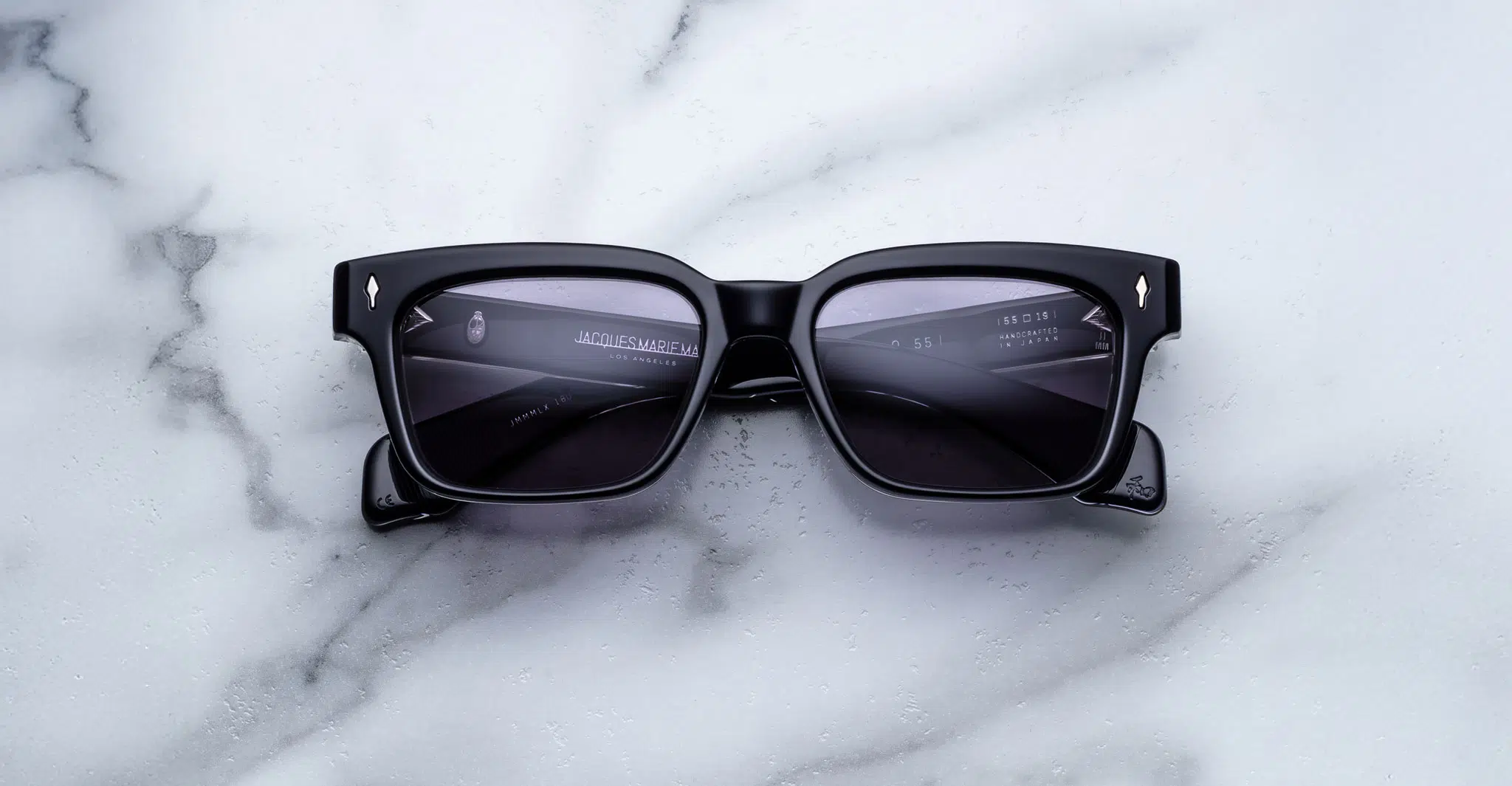 The Jacques Marie Mage Molino 55, black rectangular smart sunglasses with thick frames, rest on a white marble surface. Digital menu interface reflections are visible in their lenses.