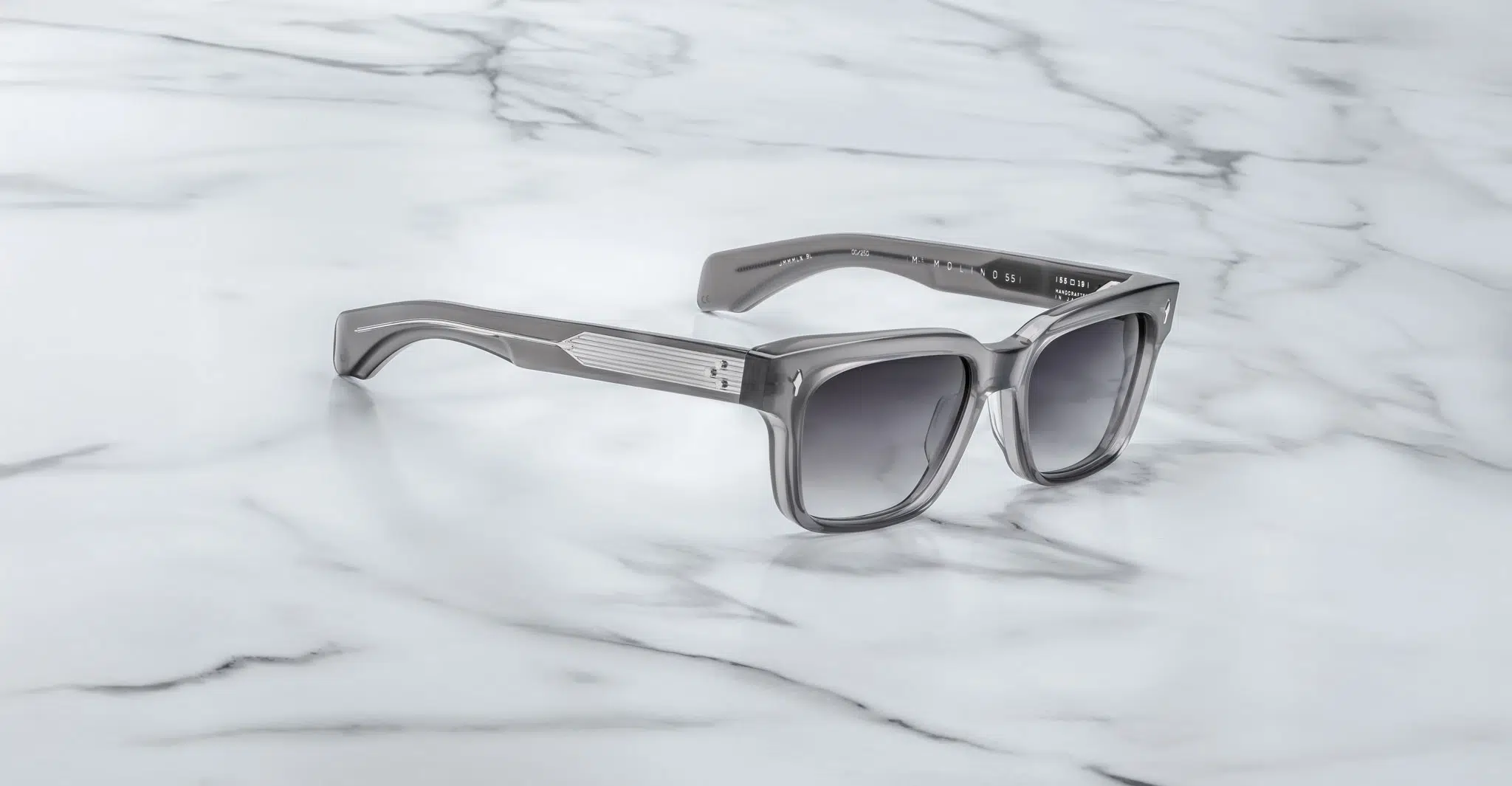 A pair of Jacques Marie Mage Molino 55 gray square-frame sunglasses with dark lenses rests on a white marble surface.