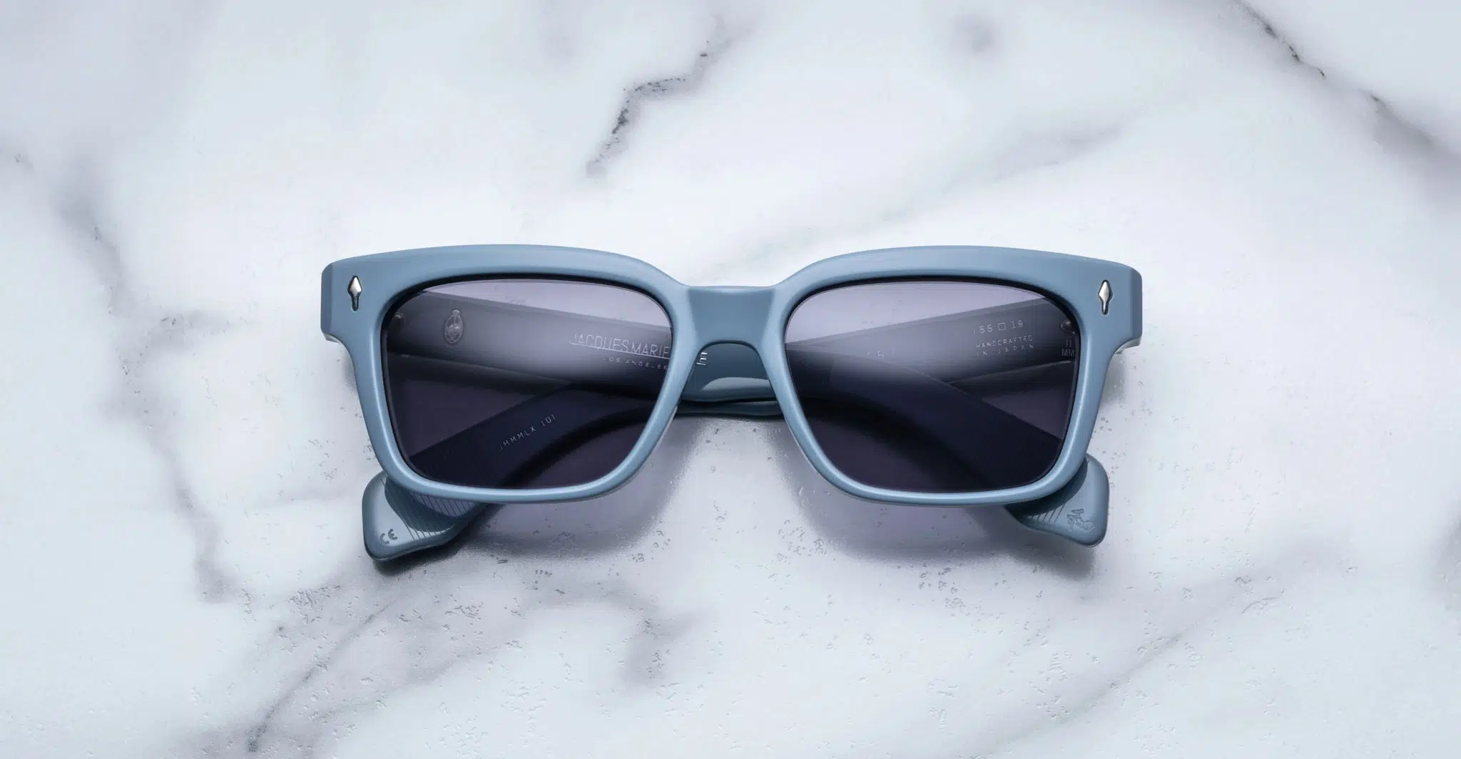 Jacques-Marie-Mage-Molino-55-Tiger-JMMMLX-10T-Front-Dan-Deutsch A pair of Jacques Marie Mage Molino 55 blue rectangular sunglasses with dark lenses rests on a white marble surface.