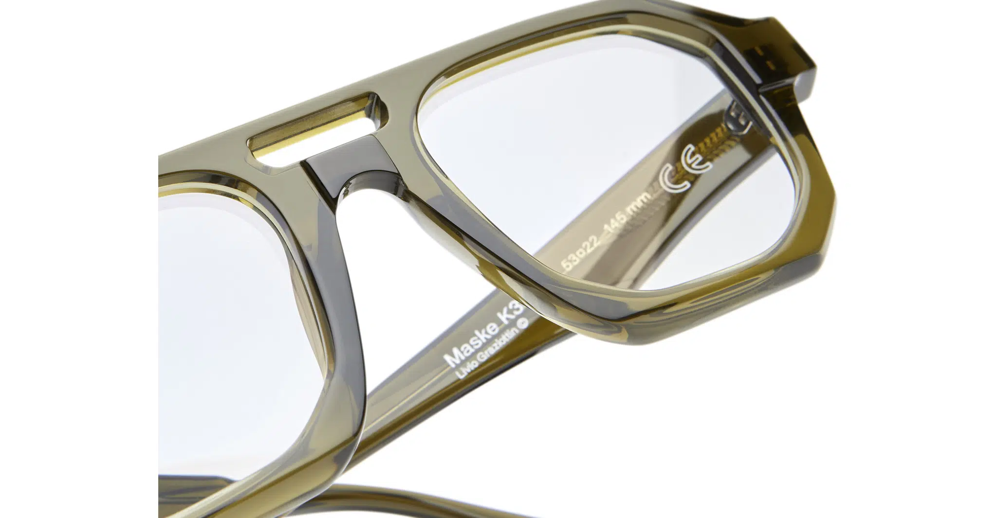 Kuboraum-K33 MO_WARM OLIVE 03-Dan-Deutsch Close-up of transparent olive-green Kuboraum K33 eyeglass frames with clear lenses, highlighting frame details, lens markings, and product info on the inner temple.