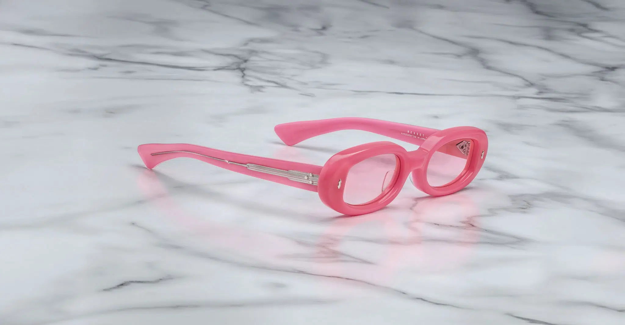 The Jacques Marie Mage Besset sunglasses, featuring round pink frames and tinted lenses, rest on a white marble surface with grey veining.