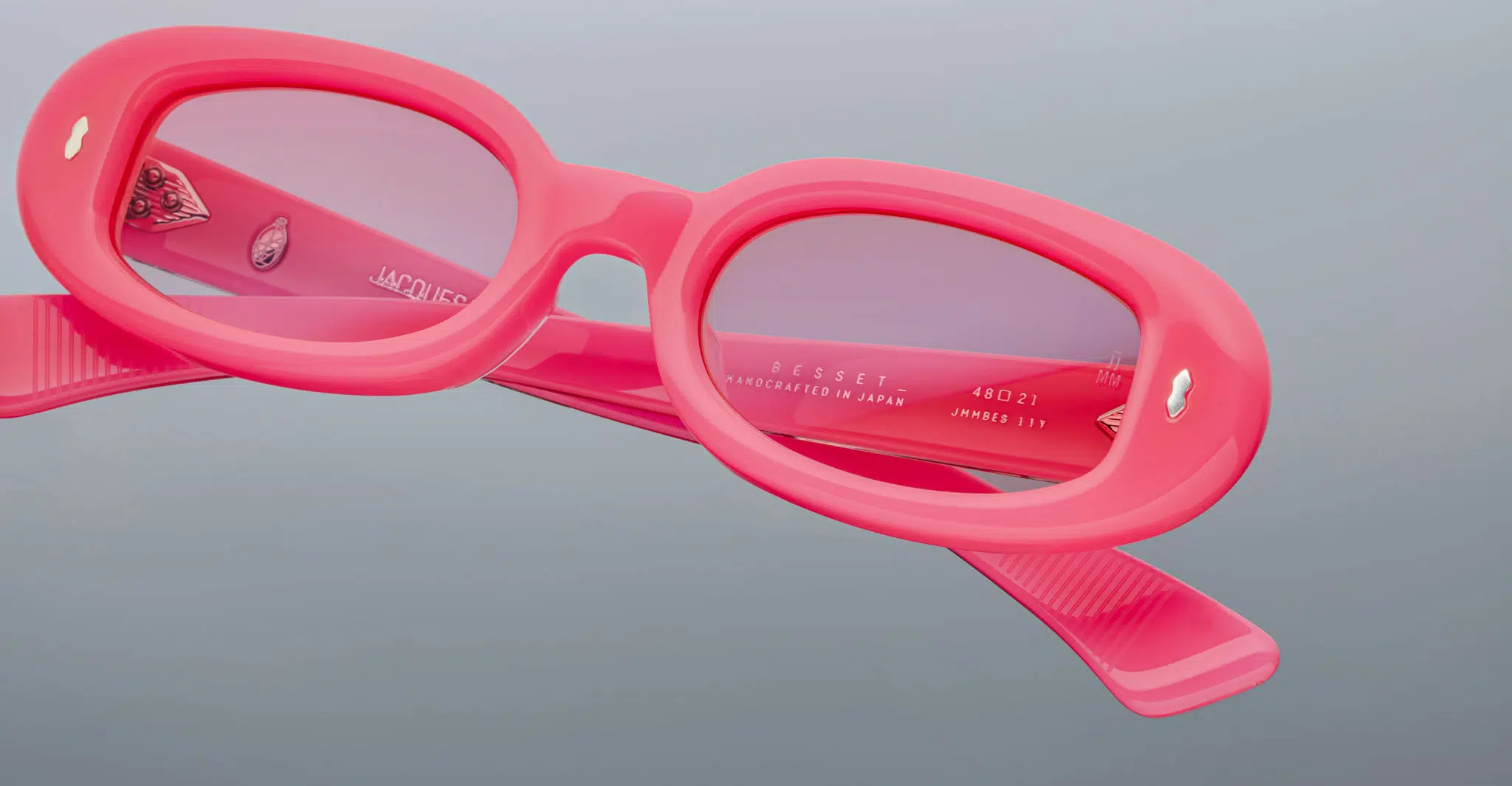 A close-up of Jacques Marie Mage Besset pink oval sunglasses with pink tinted lenses, featuring an engraved inside arm displaying the model and manufacturing details, set against a plain gray background.