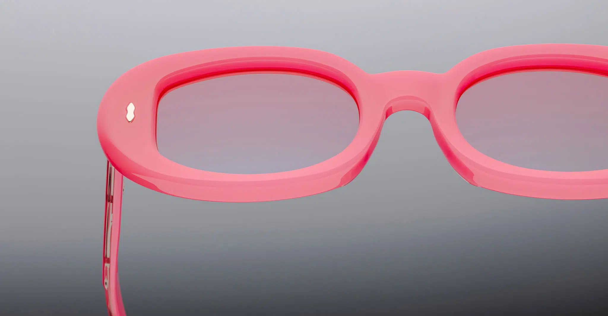 Close-up of the top part of Jacques Marie Mage Besset eyeglasses featuring thick, oval-shaped pink frames and tinted lenses set against a neutral gray background.