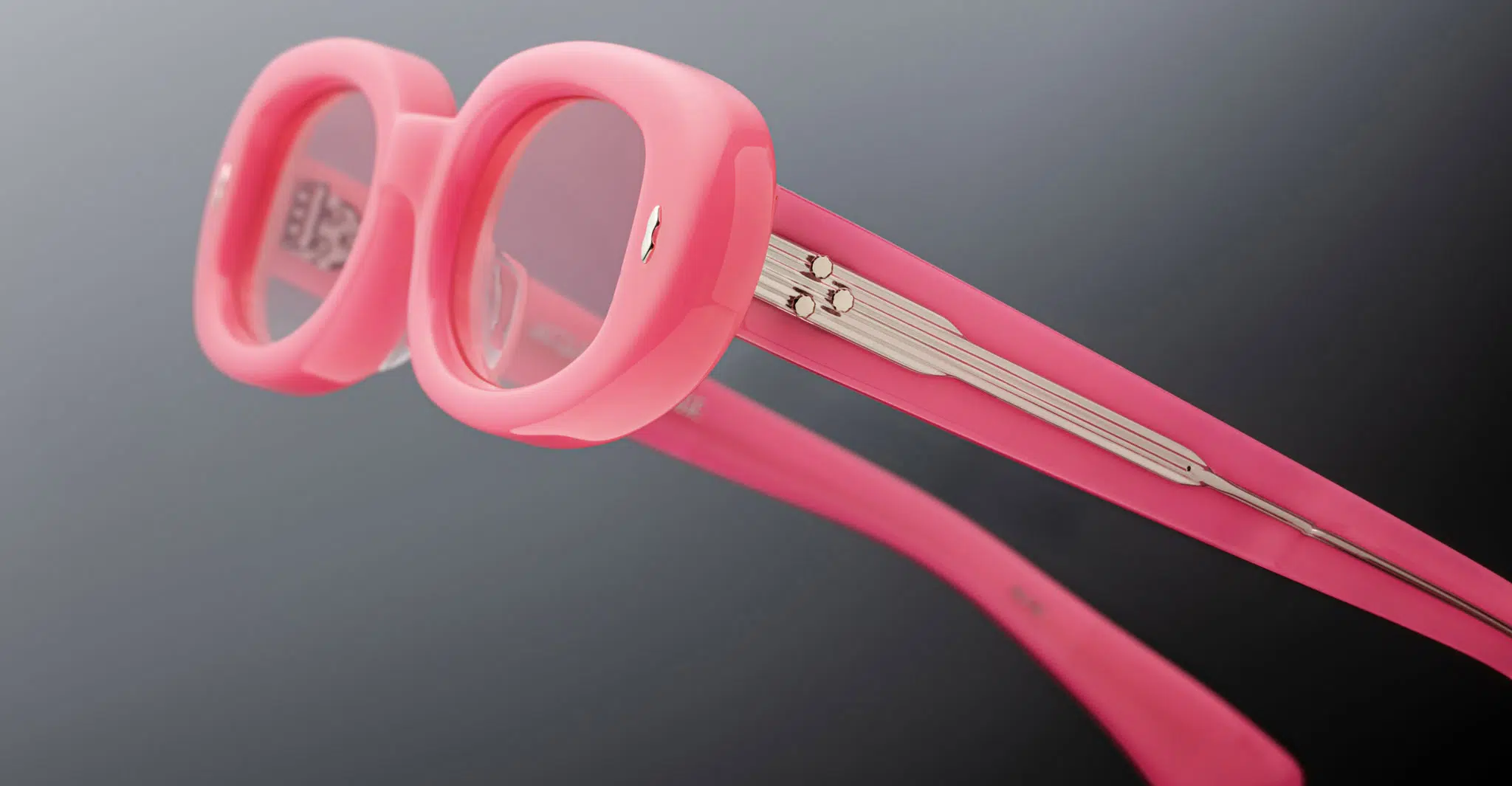 Close-up of Jacques Marie Mage Besset eyeglasses featuring thick, rounded pink frames and matching temples with visible metal hinges adorned with a linear design. The background is a soft, blurred gray.