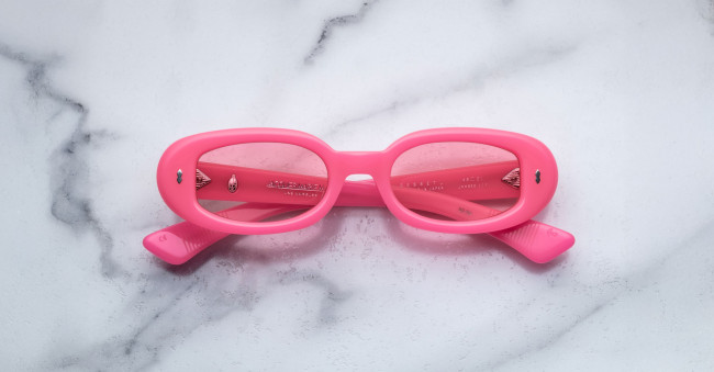 A pair of Jacques Marie Mage Besset bright pink oval sunglasses with tinted lenses is placed upside down on a white marble surface.