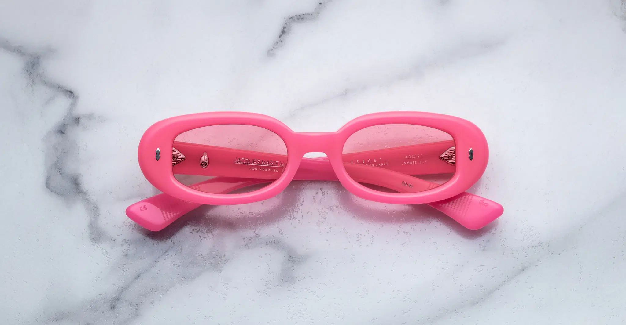 A pair of Jacques Marie Mage Besset bright pink oval sunglasses with tinted lenses is placed upside down on a white marble surface.