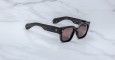 A pair of Jacques Marie Mage Enzo black rectangular sunglasses with dark lenses, thick frames, and subtle silver accents on the front, resting on a light marble surface.