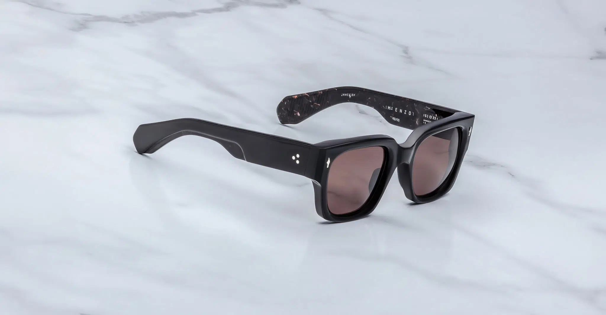 A pair of Jacques Marie Mage Enzo black rectangular sunglasses with dark lenses, thick frames, and subtle silver accents on the front, resting on a light marble surface.