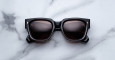 The Jacques Marie Mage Enzo, square-shaped black sunglasses with dark lenses, are displayed on a white marble surface with gray veining.
