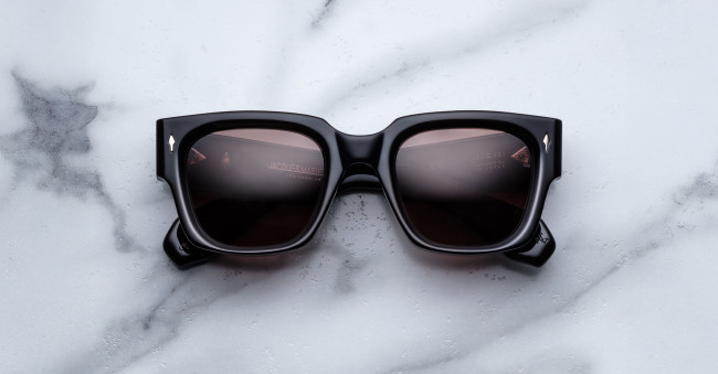 The Jacques Marie Mage Enzo, square-shaped black sunglasses with dark lenses, are displayed on a white marble surface with gray veining.