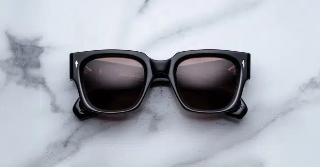 The Jacques Marie Mage Enzo, square-shaped black sunglasses with dark lenses, are displayed on a white marble surface with gray veining.