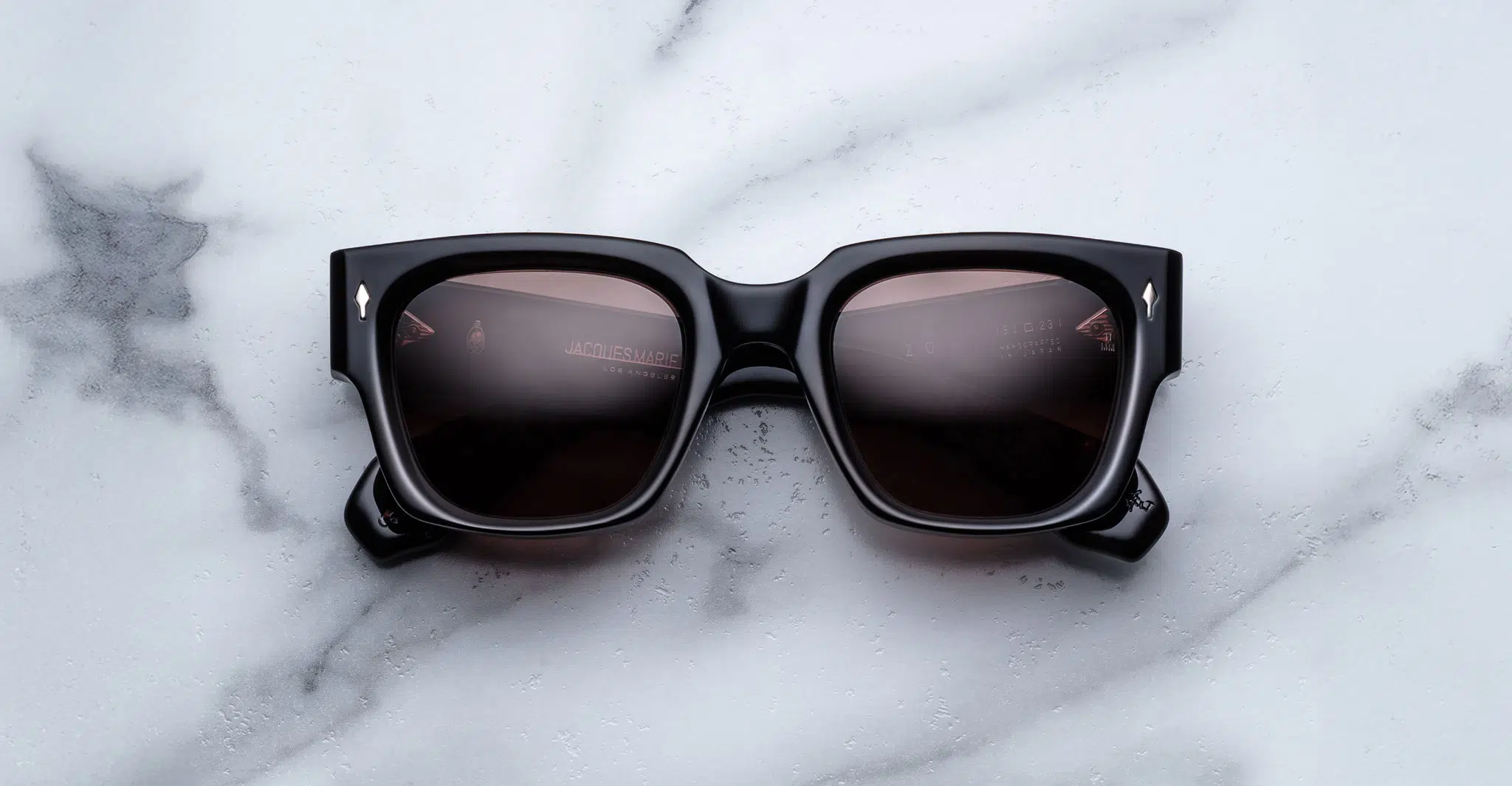 The Jacques Marie Mage Enzo, square-shaped black sunglasses with dark lenses, are displayed on a white marble surface with gray veining.