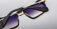 Close-up of two Jacques Marie Mage GT-R sunglasses with gold frames, black arms, and gradient purple lenses, one stacked atop the other against a plain grey background.