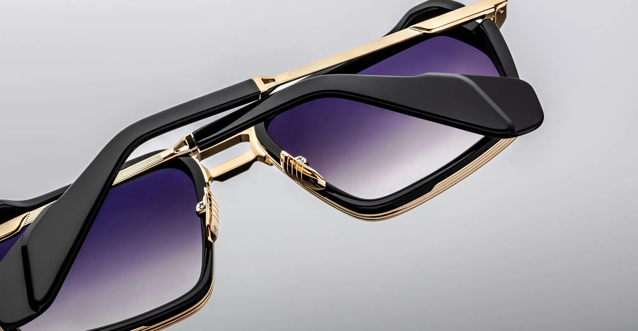 Close-up of two Jacques Marie Mage GT-R sunglasses with gold frames, black arms, and gradient purple lenses, one stacked atop the other against a plain grey background.