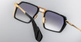 A close-up view of Jacques Marie Mage GT-R square-shaped sunglasses featuring gold metal frames, black accents, and dark gradient lenses set against a light gray background.