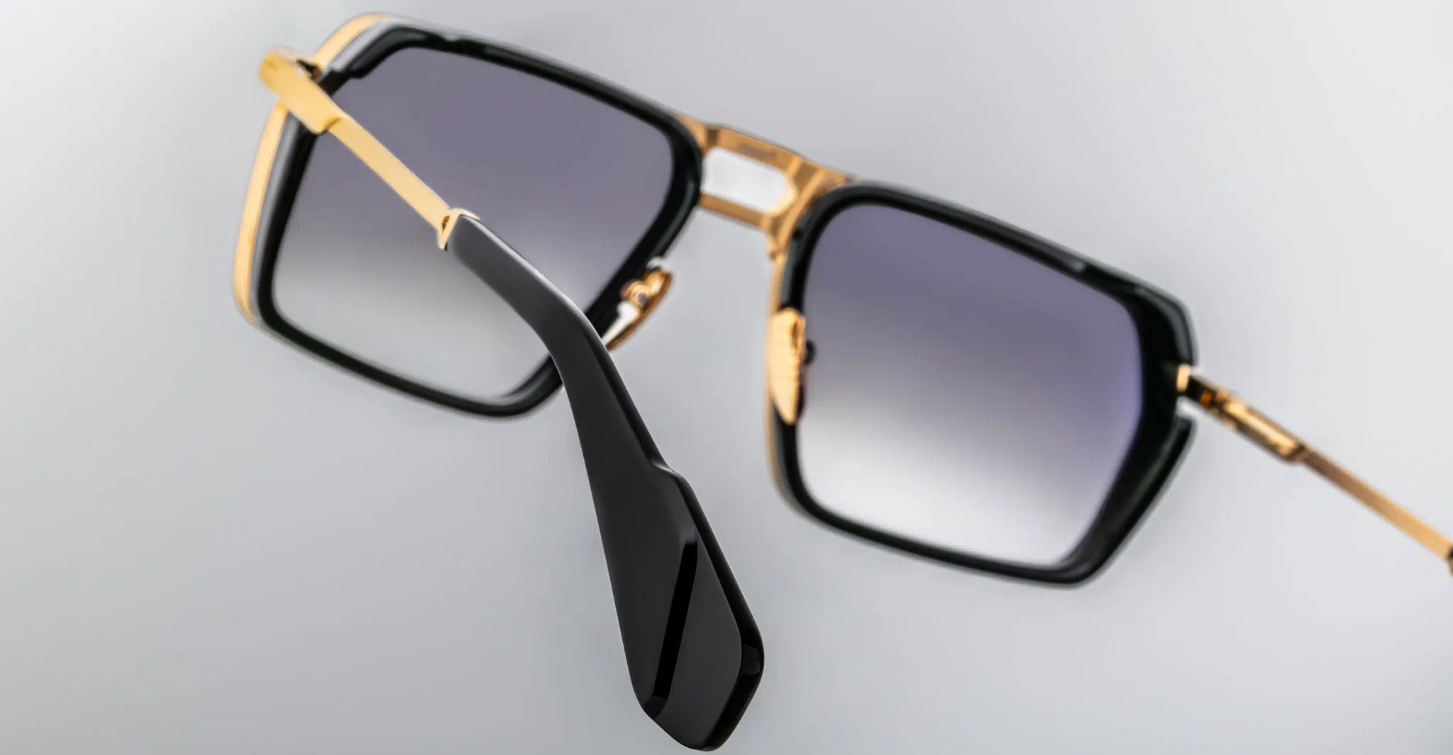 A close-up view of Jacques Marie Mage GT-R square-shaped sunglasses featuring gold metal frames, black accents, and dark gradient lenses set against a light gray background.