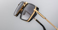 Close-up of Jacques Marie Mage GT-R eyeglasses featuring black square frames, gold accents, and dark-tinted lenses displayed against a plain light gray background.