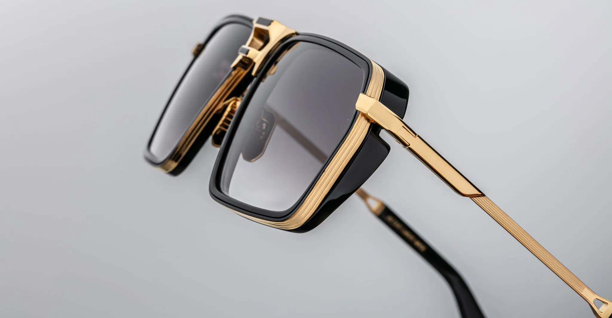 Close-up of Jacques Marie Mage GT-R eyeglasses featuring black square frames, gold accents, and dark-tinted lenses displayed against a plain light gray background.