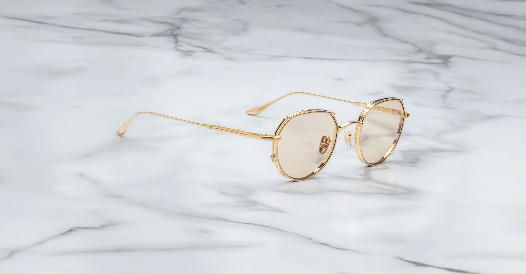 Jacques Marie Mage Hartana in Dorset JMMTN-12Q Jacques Marie Mage Hartana gold-framed round sunglasses with lightly tinted lenses rest on a smooth white marble surface with gray veining.