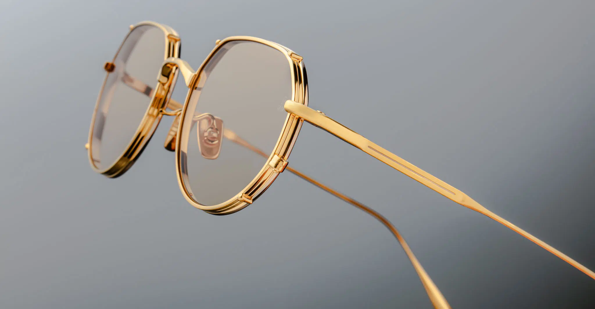 Jacques Marie Mage Hartana in Dorset JMMTN-12Q Close-up of Jacques Marie Mage Hartana eyeglasses featuring stylish gold frames, round lenses with a light brown tint, and thin temples, set against a plain grey background.