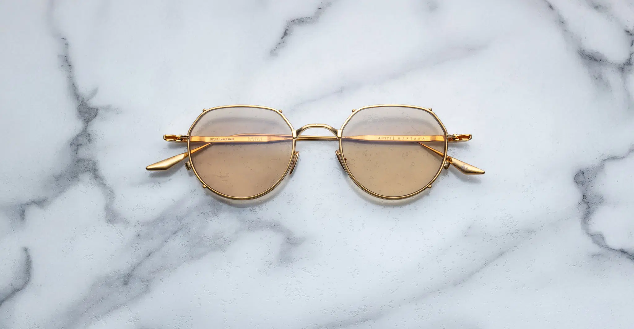 Jacques Marie Mage Hartana in Dorset JMMTN-12Q The Jacques Marie Mage Hartana features gold frames and round brown-tinted lenses, displayed on a white marble surface with grey veining.