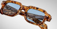 Close-up of Jacques Marie Mage Sandro eyeglasses with a thick, square tortoiseshell frame and blue-tinted lenses, resting against another pair with a translucent, textured frame.