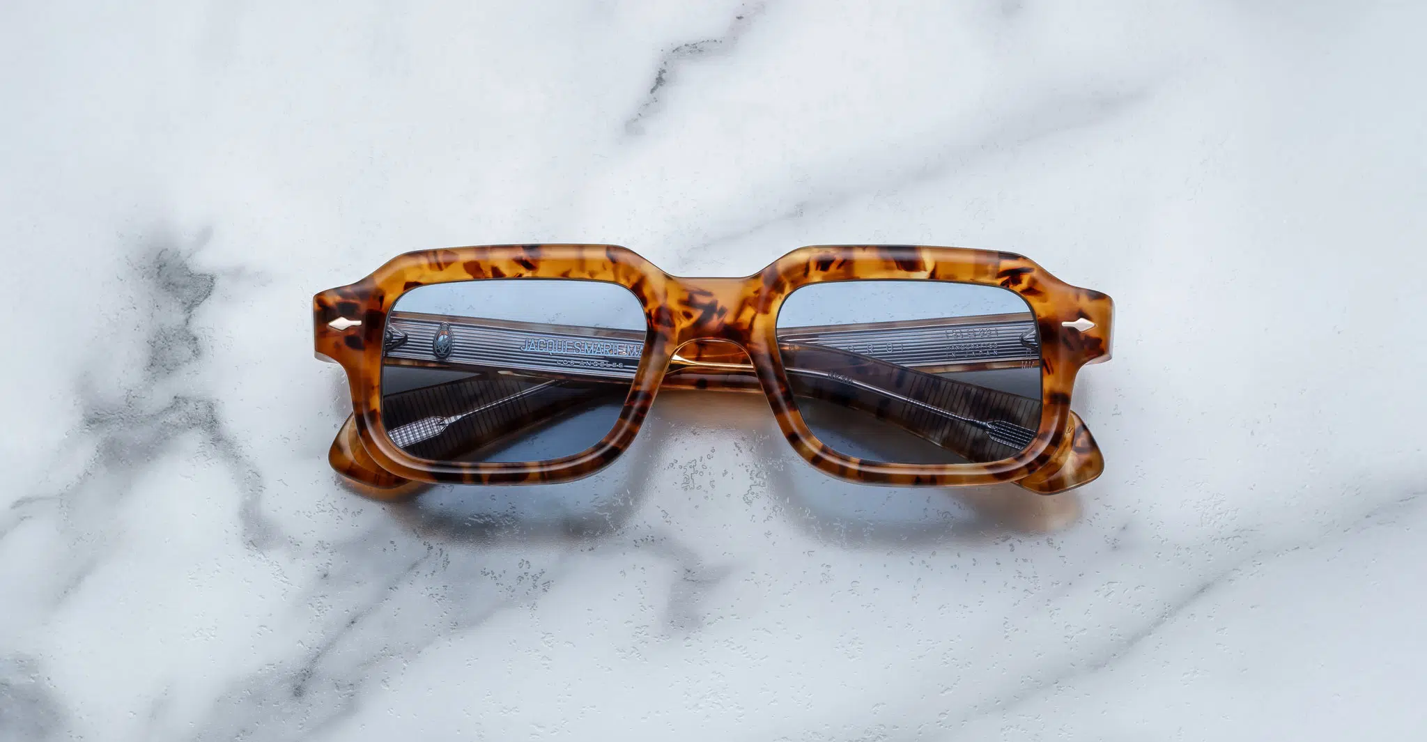 A pair of Jacques Marie Mage Sandro square tortoiseshell sunglasses with blue-tinted lenses rests on a white marble surface.