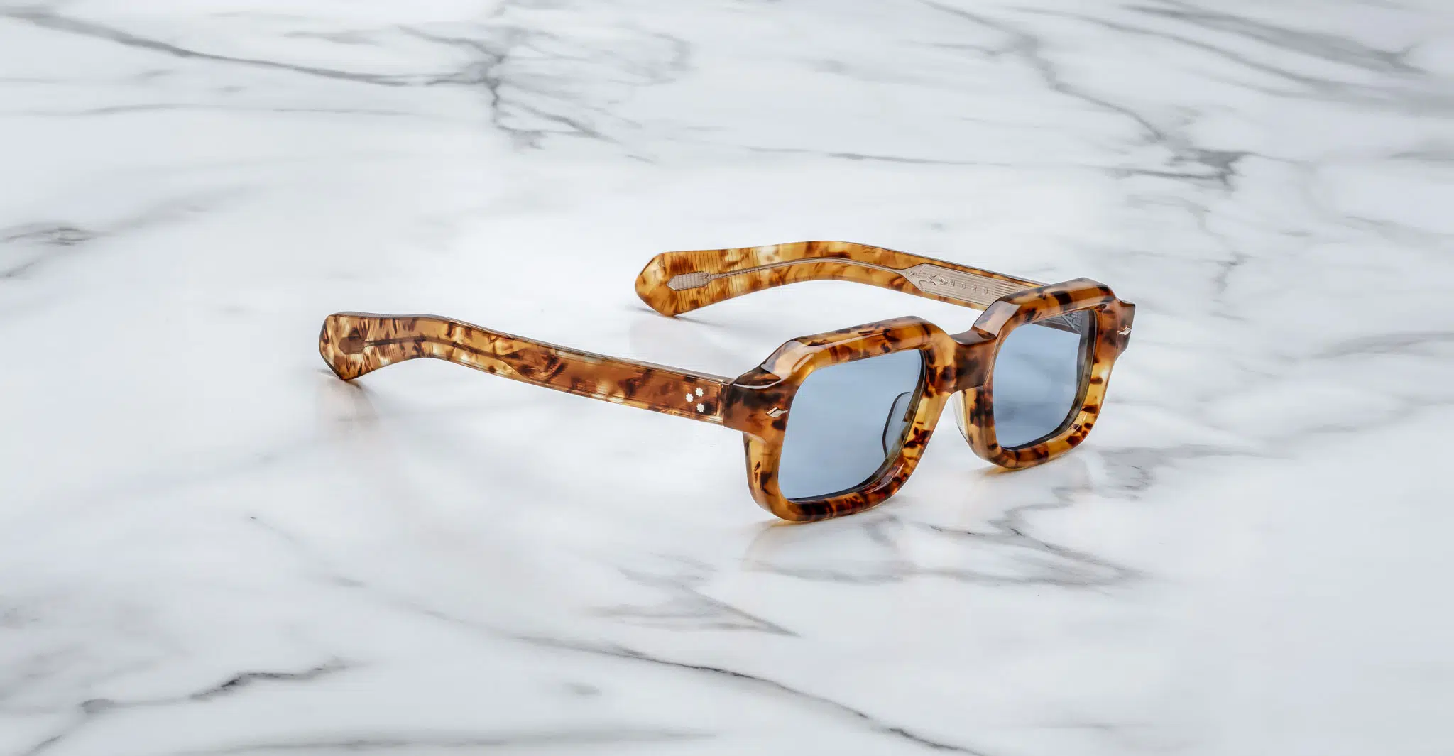 A pair of Jacques Marie Mage Sandro tortoiseshell square sunglasses with blue-tinted lenses rests on a white marble surface.