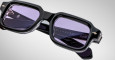 Close-up of Jacques Marie Mage Sandro black rectangular eyeglass frames with purple-tinted lenses. Sandro and Handmade in Japan are engraved inside the arms. Set against a plain gray background.