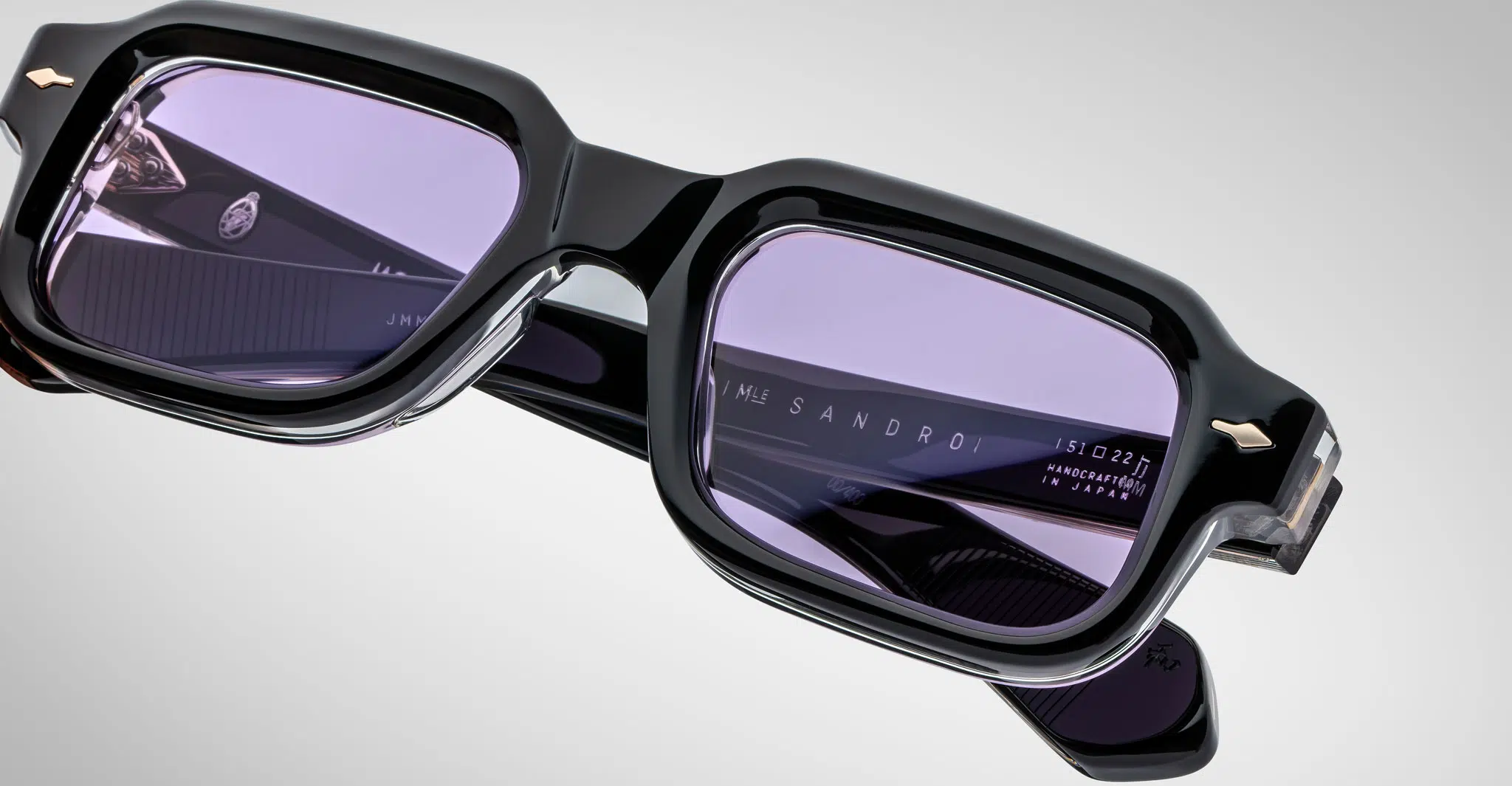 Close-up of Jacques Marie Mage Sandro black rectangular eyeglass frames with purple-tinted lenses. Sandro and Handmade in Japan are engraved inside the arms. Set against a plain gray background.