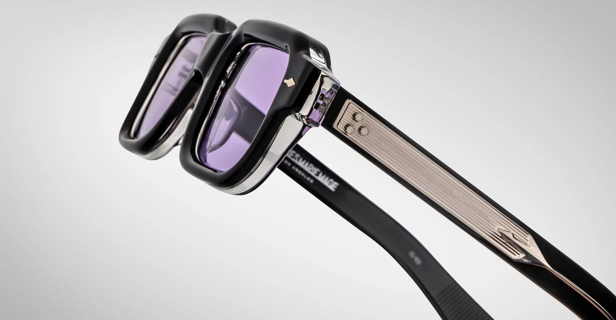 Close-up of Jacques Marie Mage Sandro sunglasses featuring thick black and silver frames, purple-tinted lenses, and textured metallic arms with decorative accents against a plain light background.
