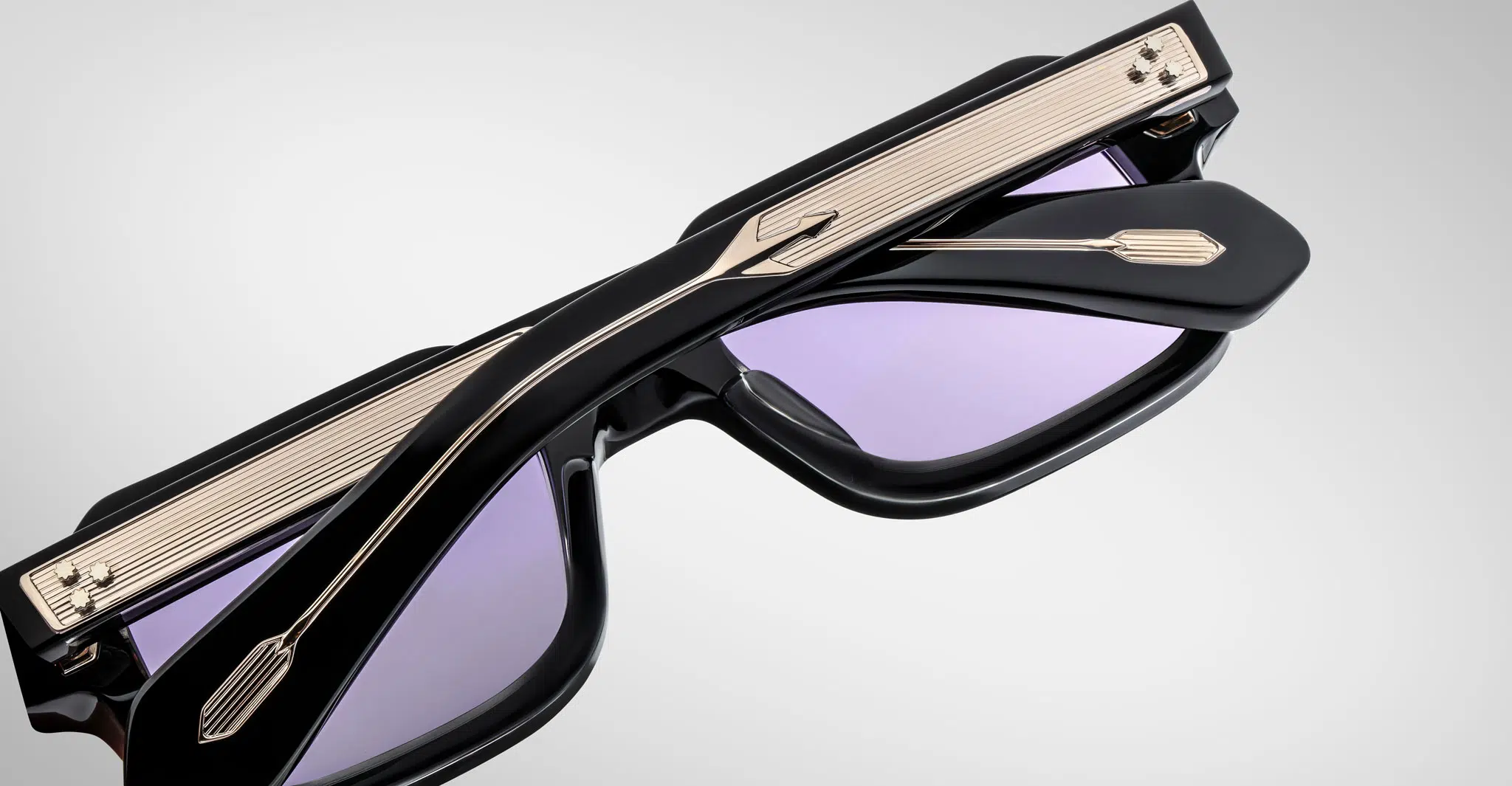 The Jacques Marie Mage Sandro sunglasses, shown folded on a light gray background, feature black frames, purple-tinted lenses, and arms with metallic grooved detailing and arrow-shaped accents.