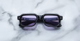A pair of Jacques Marie Mage Sandro black rectangular sunglasses with purple-tinted lenses rests on a white and gray marble surface. Branding details are printed in small white text on the left lens.