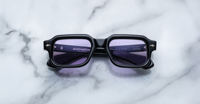 A pair of Jacques Marie Mage Sandro black rectangular sunglasses with purple-tinted lenses rests on a white and gray marble surface. Branding details are printed in small white text on the left lens.