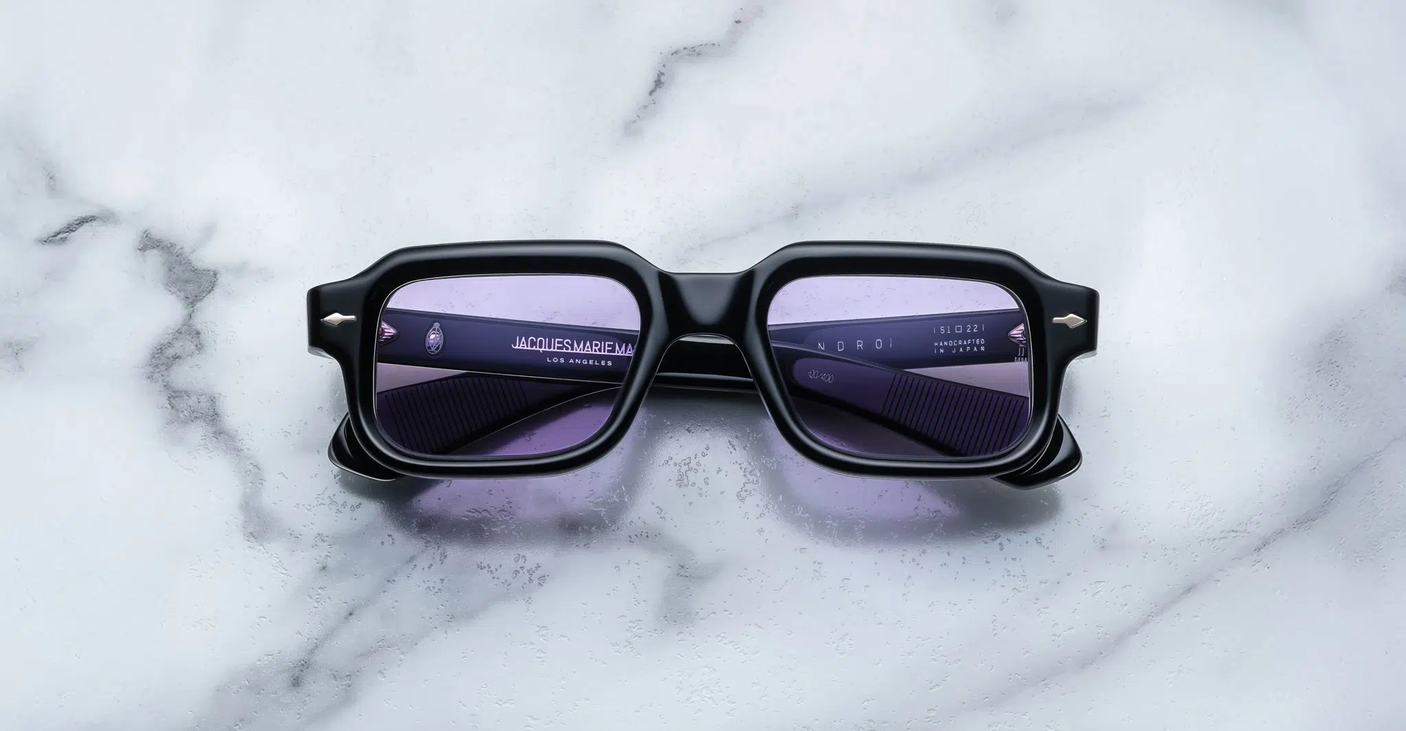 A pair of Jacques Marie Mage Sandro black rectangular sunglasses with purple-tinted lenses rests on a white and gray marble surface. Branding details are printed in small white text on the left lens.