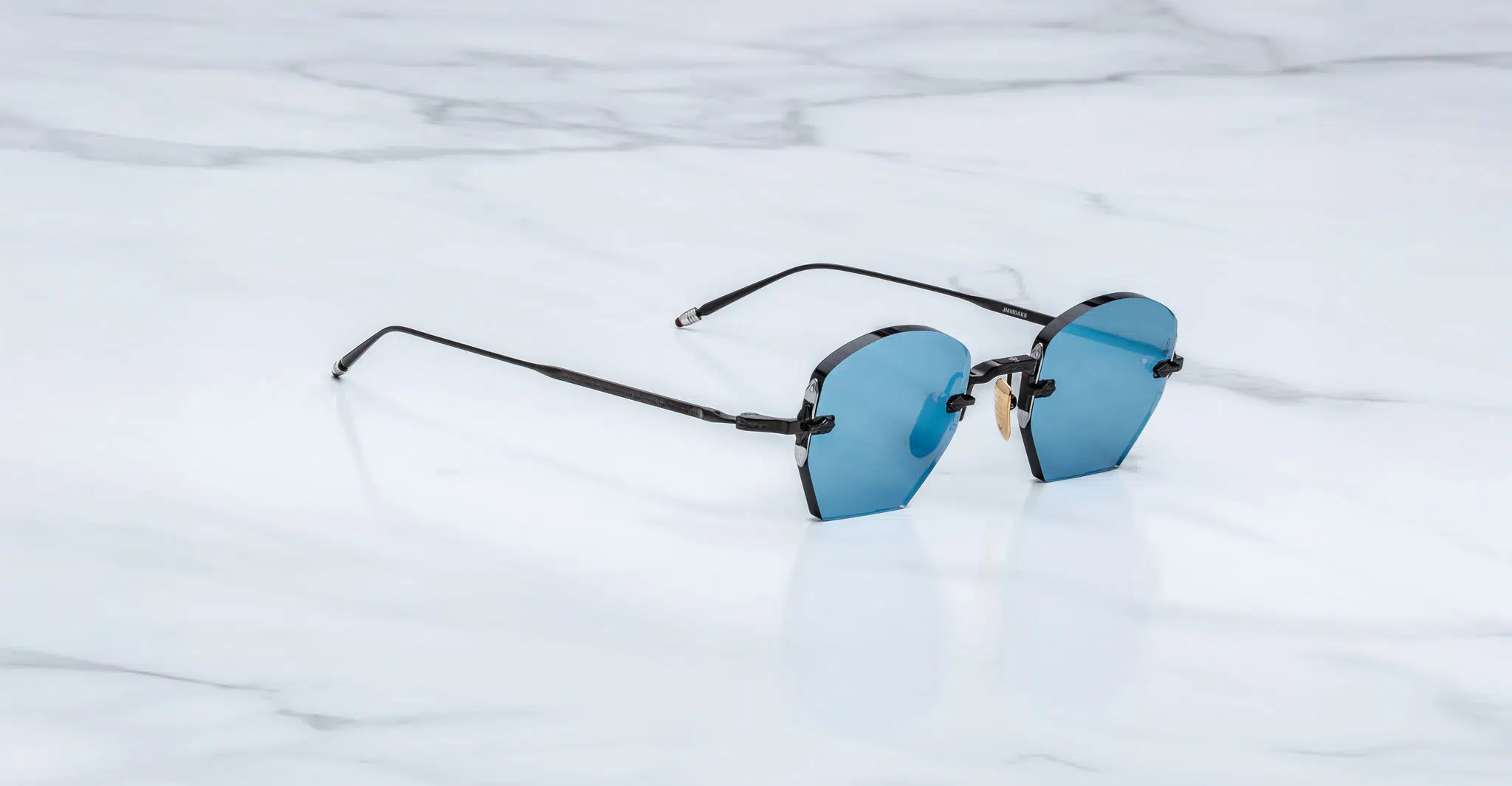 Jacques-Marie-Mage-Oatman-Lagoon-JMMOA-K8-Profile-Dan-Deutsch The Jacques Marie Mage Oatman sunglasses feature rimless blue-tinted lenses and thin black arms, displayed on a white marble surface.