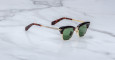 The Jacques Marie Mage Sartre sunglasses feature green lenses, gold frames, black top rim accents, and tortoiseshell-patterned arms, shown on a white marble surface.
