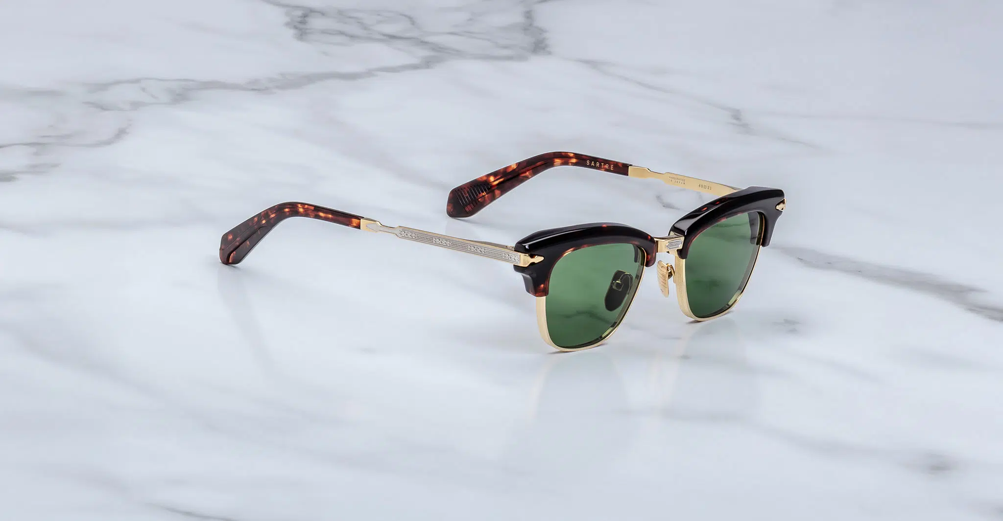 The Jacques Marie Mage Sartre sunglasses feature green lenses, gold frames, black top rim accents, and tortoiseshell-patterned arms, shown on a white marble surface.