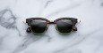 A pair of Jacques Marie Mage Sartre black and gold retro-style sunglasses with dark lenses rests on a white marble surface.