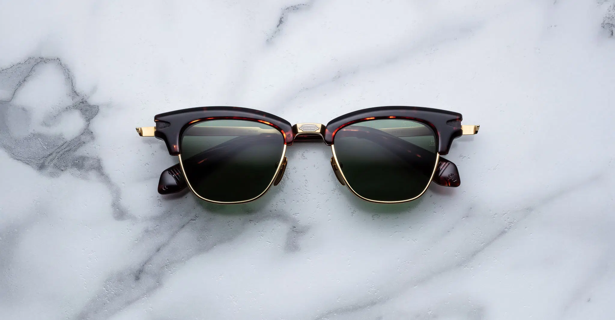 A pair of Jacques Marie Mage Sartre black and gold retro-style sunglasses with dark lenses rests on a white marble surface.