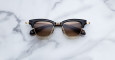 The Jacques Marie Mage Sartre sunglasses feature black and gold frames with brown tinted lenses, shown on a white marble surface.