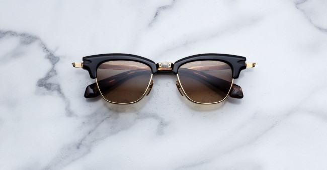The Jacques Marie Mage Sartre sunglasses feature black and gold frames with brown tinted lenses, shown on a white marble surface.