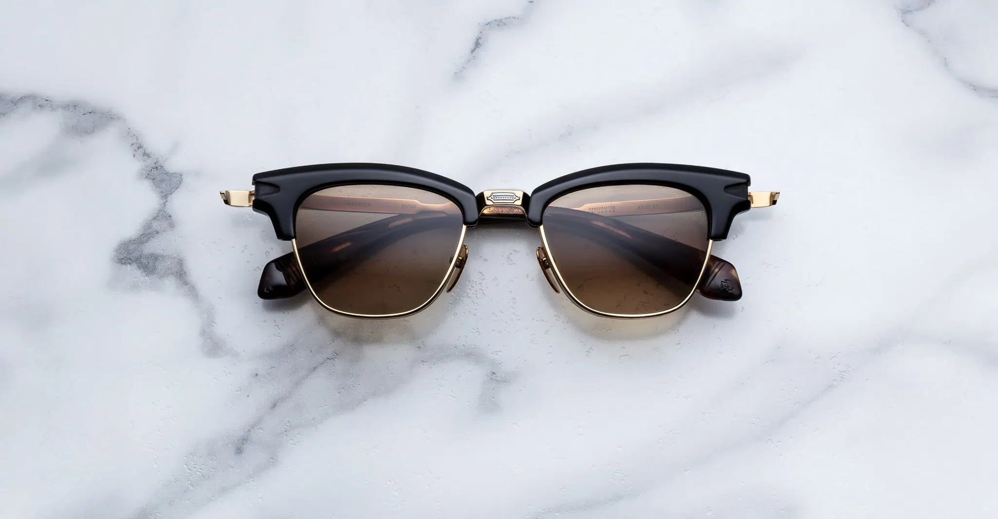 Jacques-Marie-Mage-Sartre-Noir-15-JMMTQ-15A-Front-Dan-Deutsch The Jacques Marie Mage Sartre sunglasses feature black and gold frames with brown tinted lenses, shown on a white marble surface.