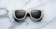 A pair of Jacques Marie Mage Chelsea beige, thick-framed sunglasses with dark lenses rests on a white marble surface with grey veins, with faint brand details visible on the inside of the left lens.