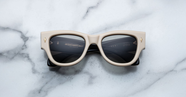 A pair of Jacques Marie Mage Chelsea beige, thick-framed sunglasses with dark lenses rests on a white marble surface with grey veins, with faint brand details visible on the inside of the left lens.